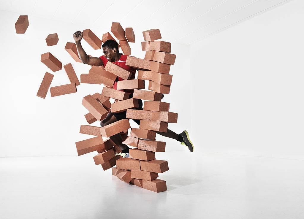 Male runner wearing black shorts and white top breaking through a brick wall.